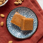 Two squares of peanut butter freezer fudge on a blue pattered plate on a red napkin next to a bowl of peanuts.