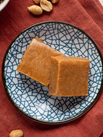 Two squares of peanut butter freezer fudge on a blue pattered plate on a red napkin next to a bowl of peanuts.