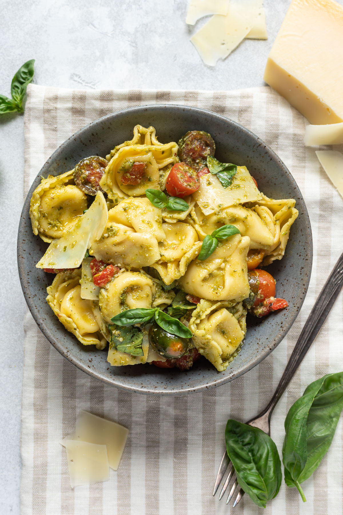 A bowl of pesto tortellini salad on a striped linen napkin next to shaved parmesan cheese and fresh basil. 
