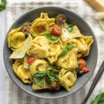 A bowl of pesto tortellini salad on a striped linen napkin next to shaved parmesan cheese and fresh basil.