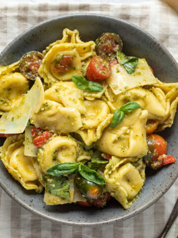 A bowl of pesto tortellini salad on a striped linen napkin next to shaved parmesan cheese and fresh basil.