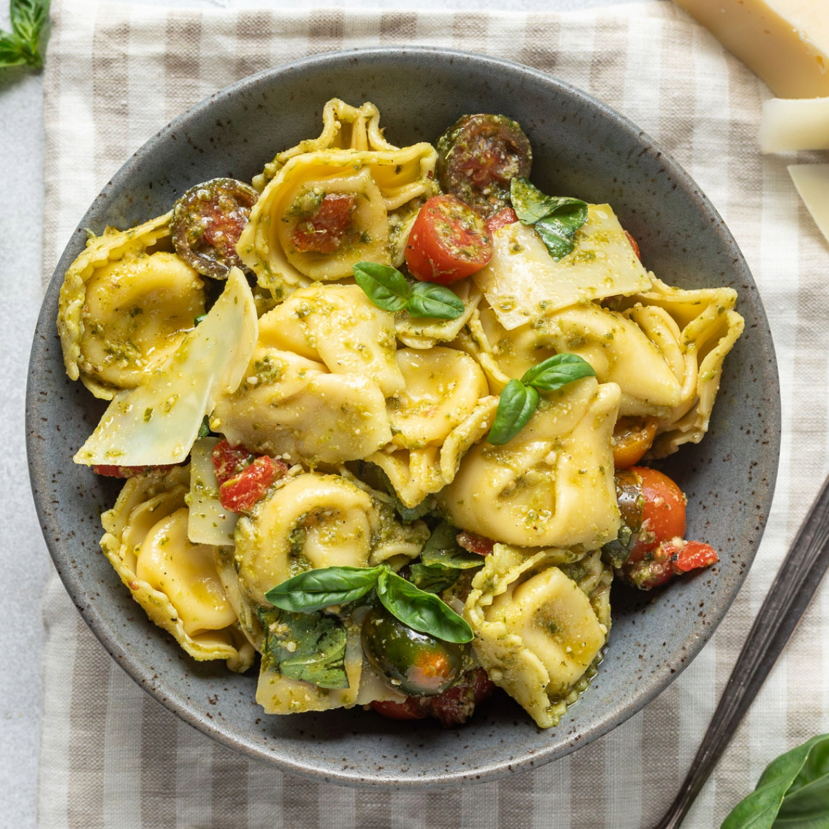 A bowl of pesto tortellini salad on a striped linen napkin next to shaved parmesan cheese and fresh basil.