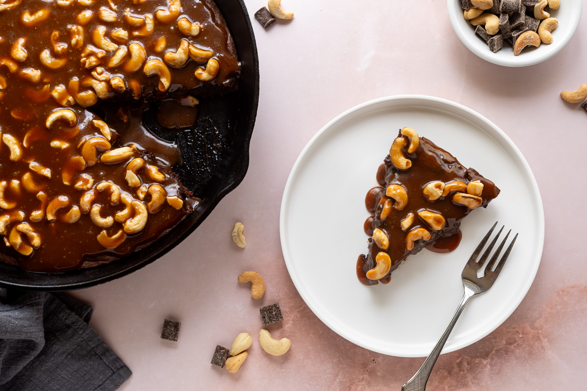 Brownie baked in a cast iron skillet topped with cashew caramel, a slice is missing from the skillet and is on a white plate next to it.
