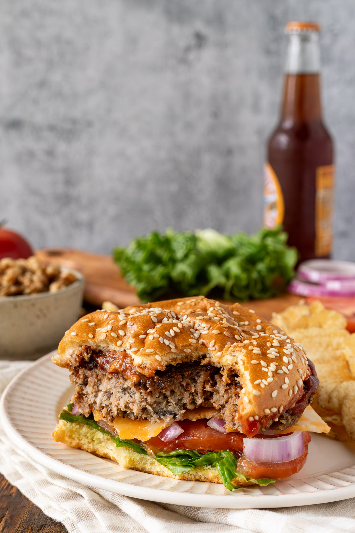 Close up image of a walnut burger with bites taken out of it. In the background you can see ingredients used to make the burger, chips, and a soda.
