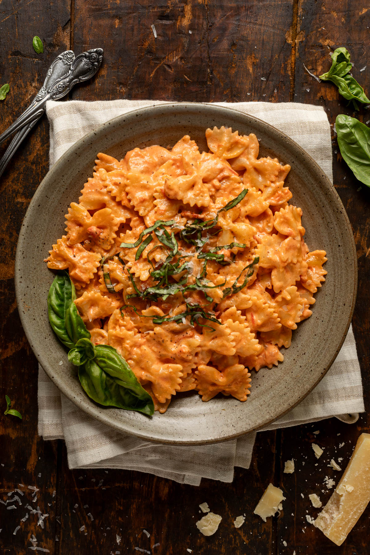 A plate of tomato alfredo pasta topped with fresh basil on a striped linen napkin. Next to the plate is a fork, more fresh basil, and a block of parmesan cheese.