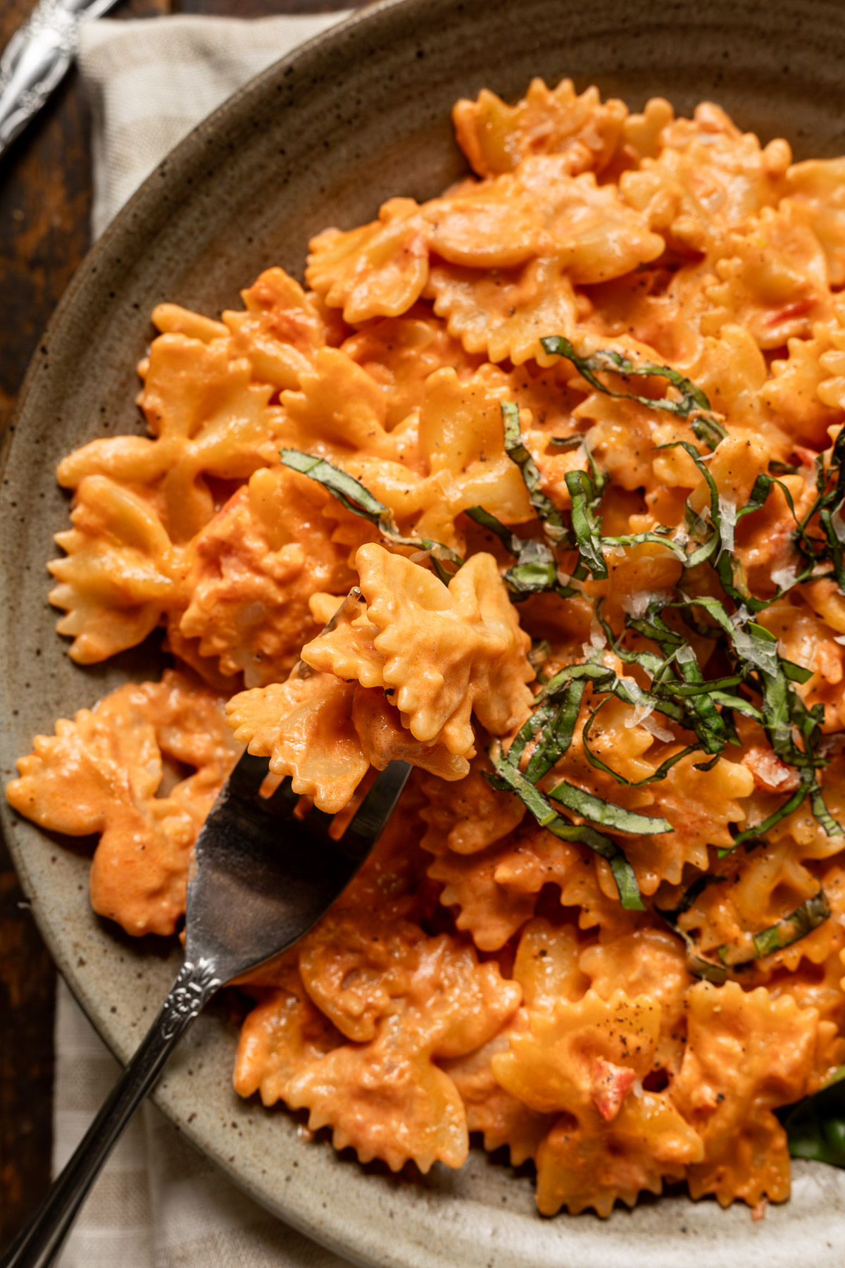 Close up image of tomato alfredo pasta on a plate topped with basil and parmesan cheese. A fork is stabbing a couple of bowtie noodles.