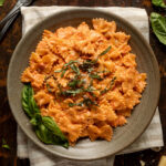 A plate of tomato alfredo pasta topped with fresh basil on a striped linen napkin. Next to the plate is a fork, more fresh basil, and a block of parmesan cheese.
