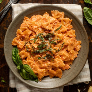 A plate of tomato alfredo pasta topped with fresh basil on a striped linen napkin. Next to the plate is a fork, more fresh basil, and a block of parmesan cheese.