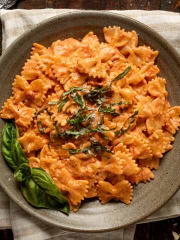 A plate of tomato alfredo pasta topped with fresh basil on a striped linen napkin. Next to the plate is a fork, more fresh basil, and a block of parmesan cheese.
