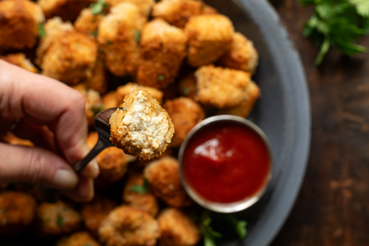 Tofu nugget on a fork bit in half, in the background you can see fried tofu nuggets on a plate garnished with parsley next to a small bowl of ketchup.