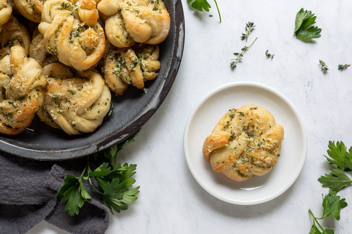 A serving platter of garlic parmesan knots next to a small plate with a single garlic knot on it.
