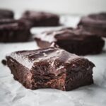 Frosted sweet potato brownies on a sheet of parchment paper, one has a bite taken out of it.