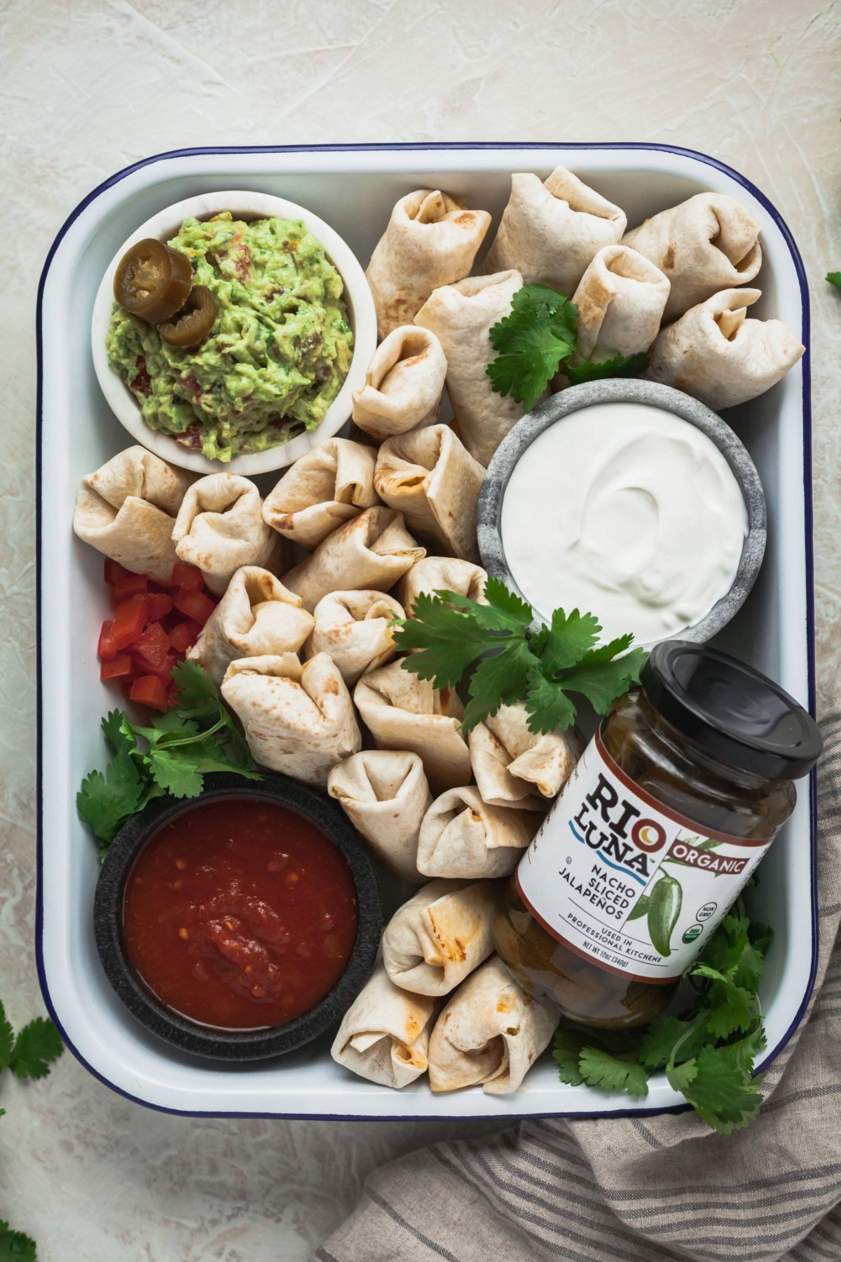Mini vegetarian burrito bites in a baking dish with small bowls of sour cream, salsa, Rio Luna peppers, and spicy guacamole.