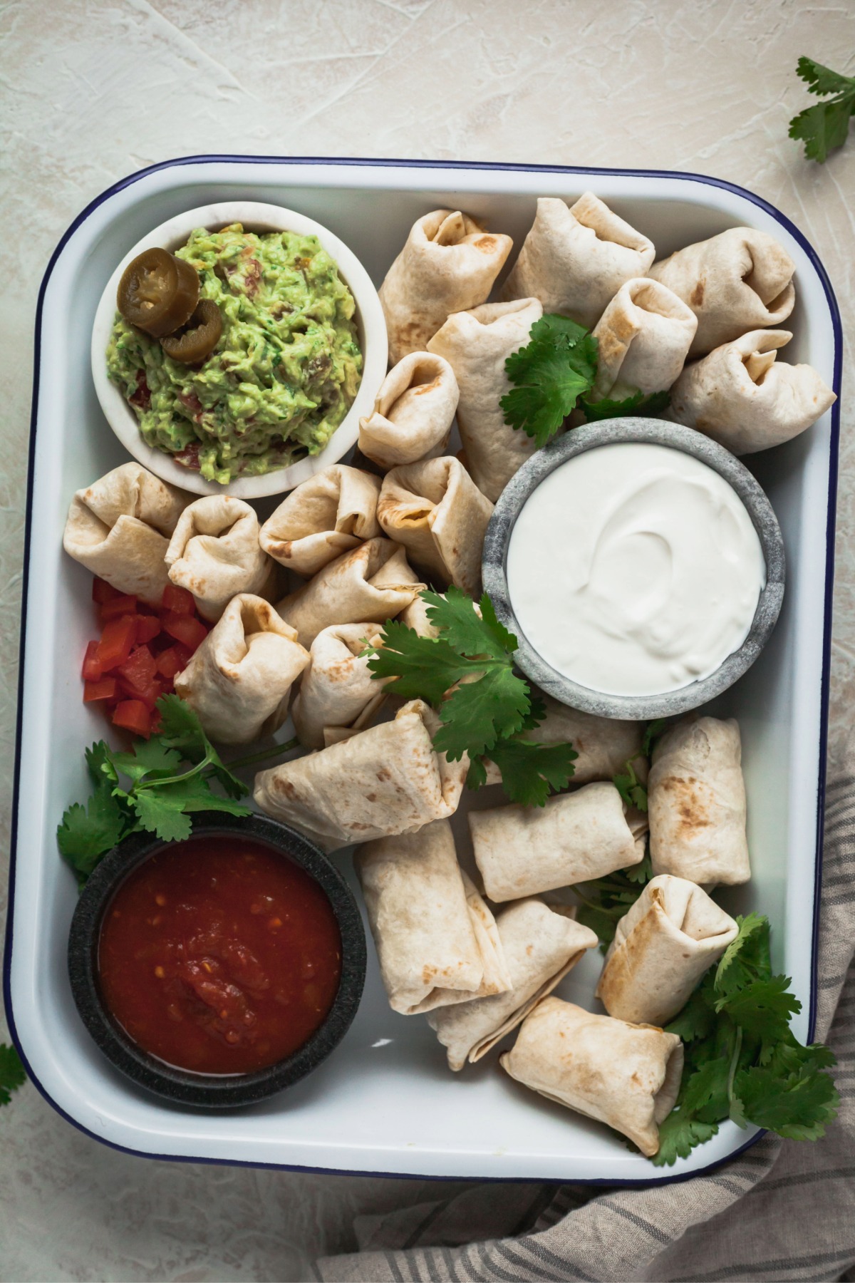 Mini vegetarian burrito bites in a baking dish with small bowls of sour cream, salsa, and spicy guacamole.