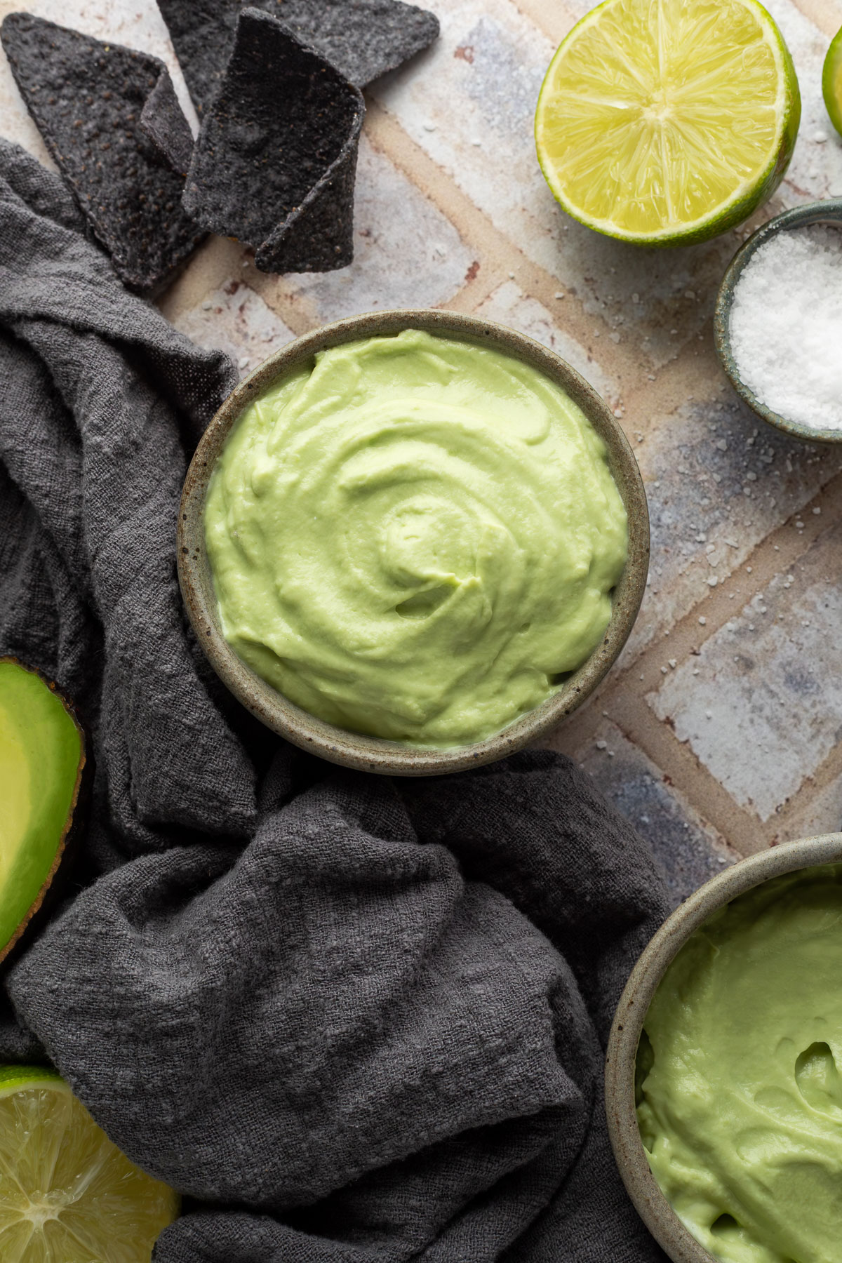 A bowl of avocado crema next to a gray linen napkin, half a lime, and a small bowl of salt.