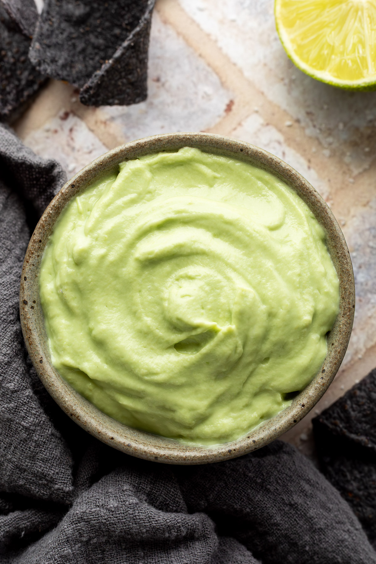 A bowl of avocado crema next to a gray linen napkin, half a lime, and a small bowl of salt.