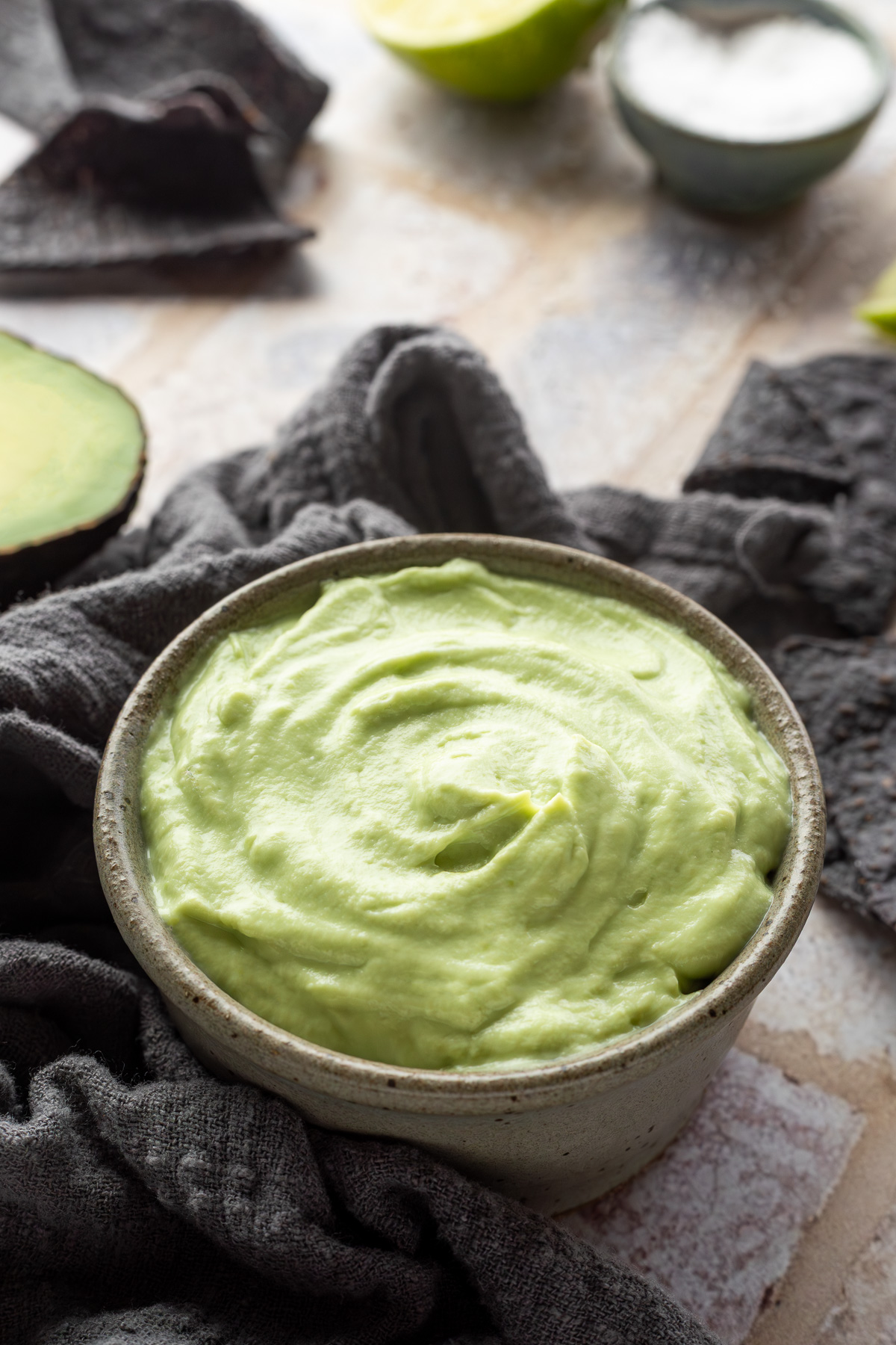 A bowl of avocado crema next to a gray linen napkin, half a lime, and a small bowl of salt.