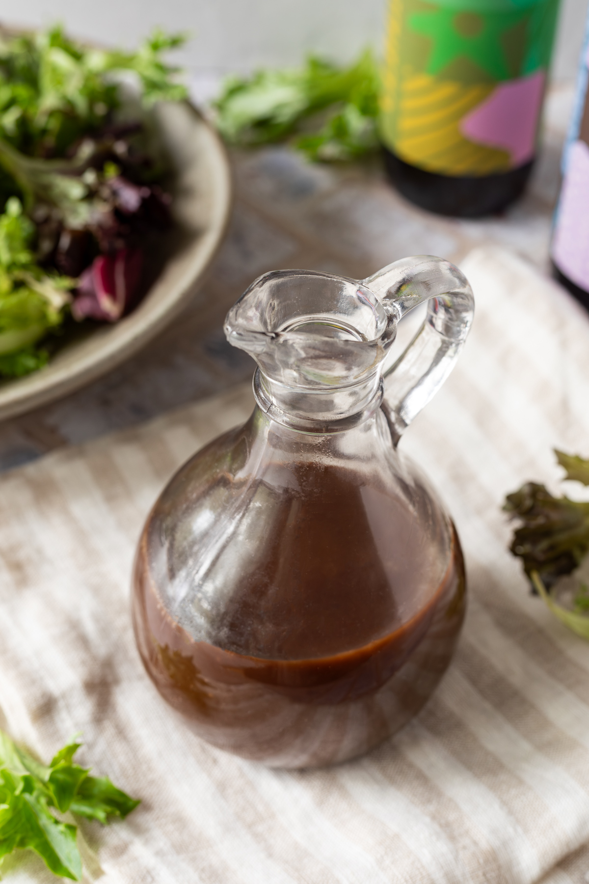 A glass jug full of balsamic vinaigrette sitting on a striped linen napkin. In the background there is a bowl of salad and two bottles of balsamic vinegar.