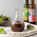 A glass jug full of balsamic vinaigrette sitting on a striped linen napkin. In the background there is a bowl of salad and two bottles of balsamic vinegar.