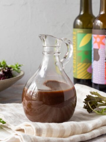 A glass jug full of balsamic vinaigrette sitting on a striped linen napkin. In the background there is a bowl of salad and two bottles of balsamic vinegar.