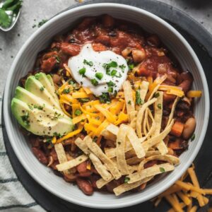 A bowl of vegetarian chili topped with sour cream, tortilla strips, sliced avocado, and fresh chives.