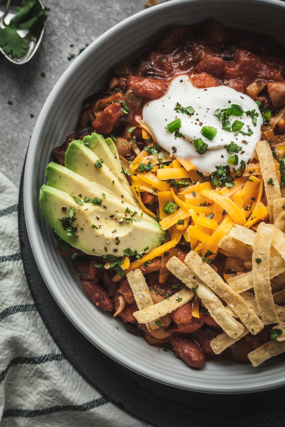 A bowl of vegetarian chili topped with sour cream, tortilla strips, sliced avocado, and fresh chives.