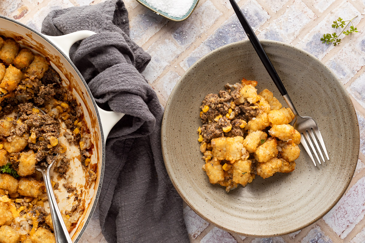 A scoop of tater tot casserole in a bowl with a fork sticking out of it next to a pan of tater tot casserole.