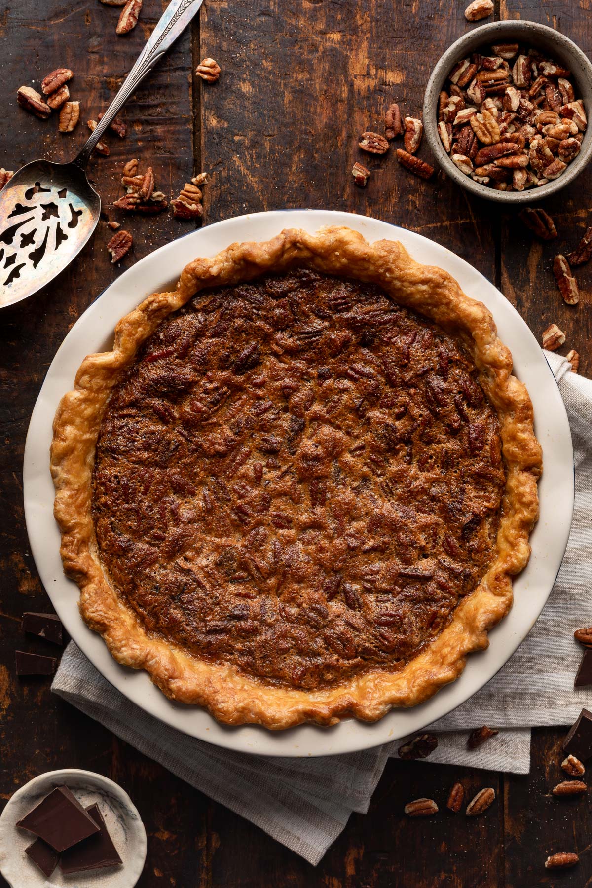 chocolate bourbon pecan pie on a wood table with a pie server, bowl of pecans, and plate of chocolate bars.