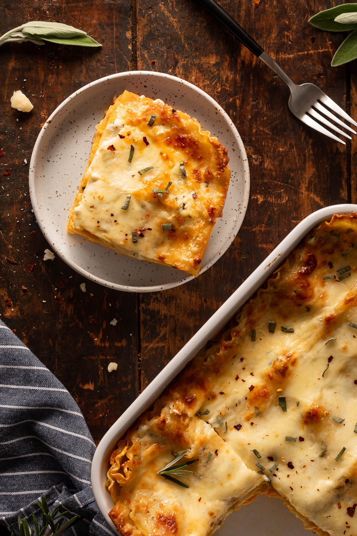 A slice of butternut squash lasagna on a plate next to a fork with the pan of butternut squash lasagna in the background.