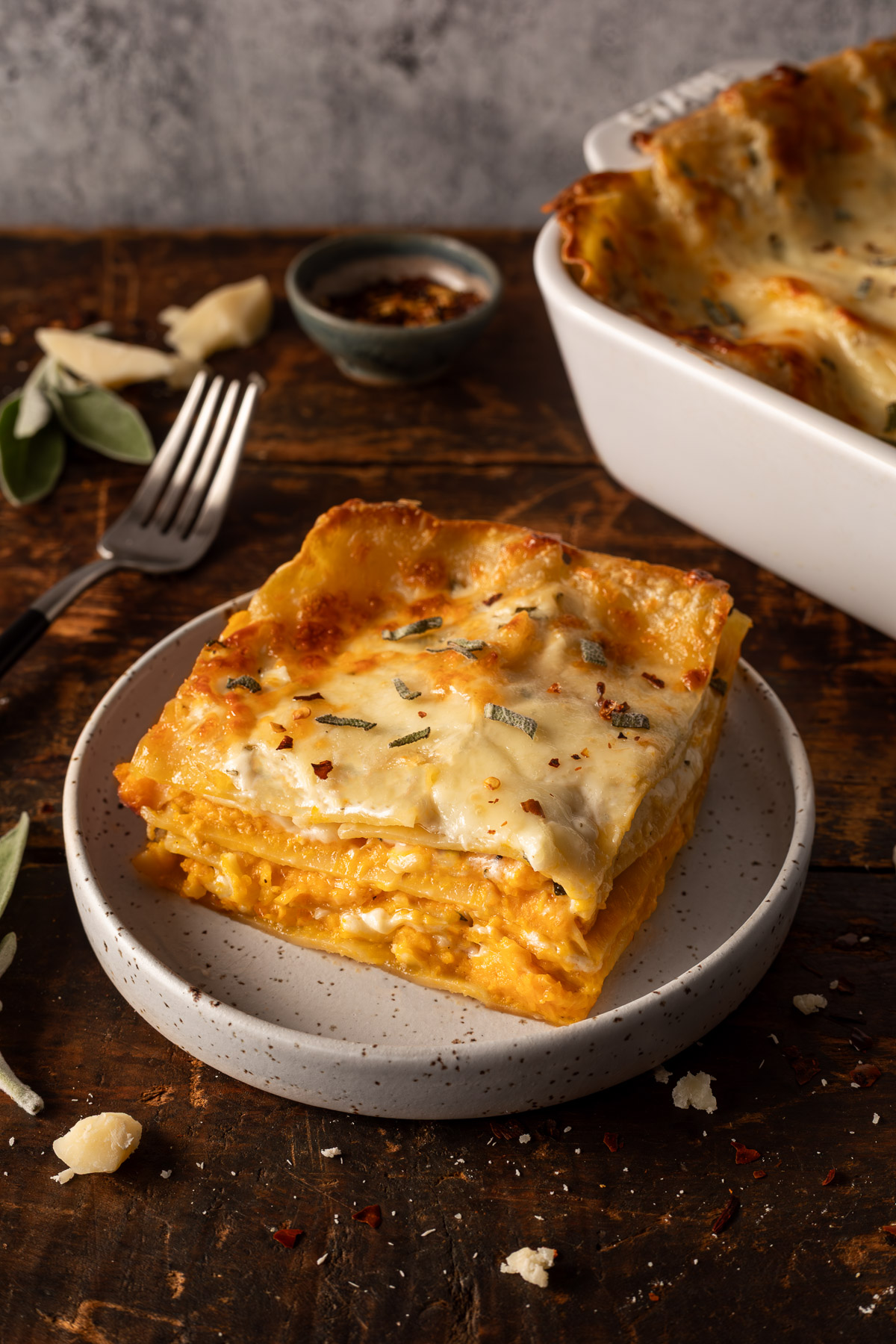 A slice of butternut squash lasagna on a plate next to a fork with the pan of butternut squash lasagna in the background.