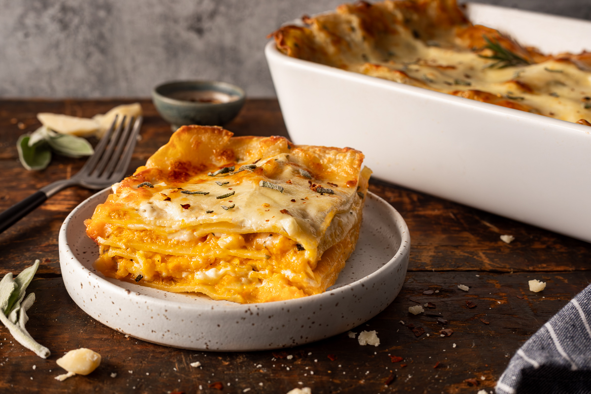 A slice of butternut squash lasagna on a plate next to a fork with the pan of butternut squash lasagna in the background.
