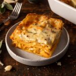 A slice of butternut squash lasagna on a plate next to a fork with the pan of butternut squash lasagna in the background.