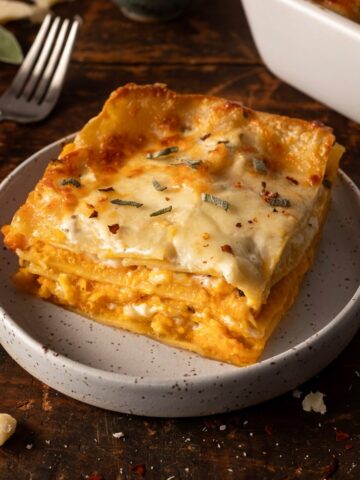 A slice of butternut squash lasagna on a plate next to a fork with the pan of butternut squash lasagna in the background.