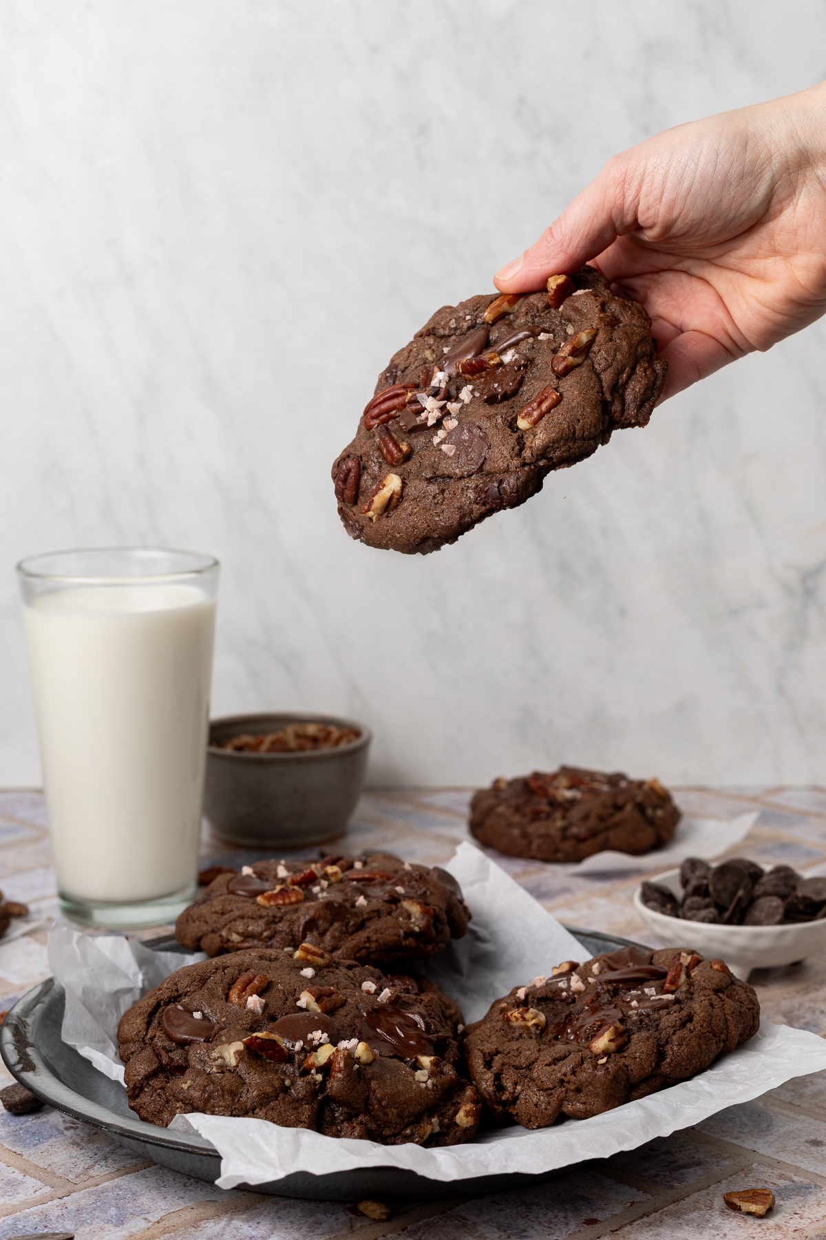 A hand holding a double chocolate chip cookie above a platter of cookies. In the background there is a tall glass of milk and a bowl of chocolate chips.