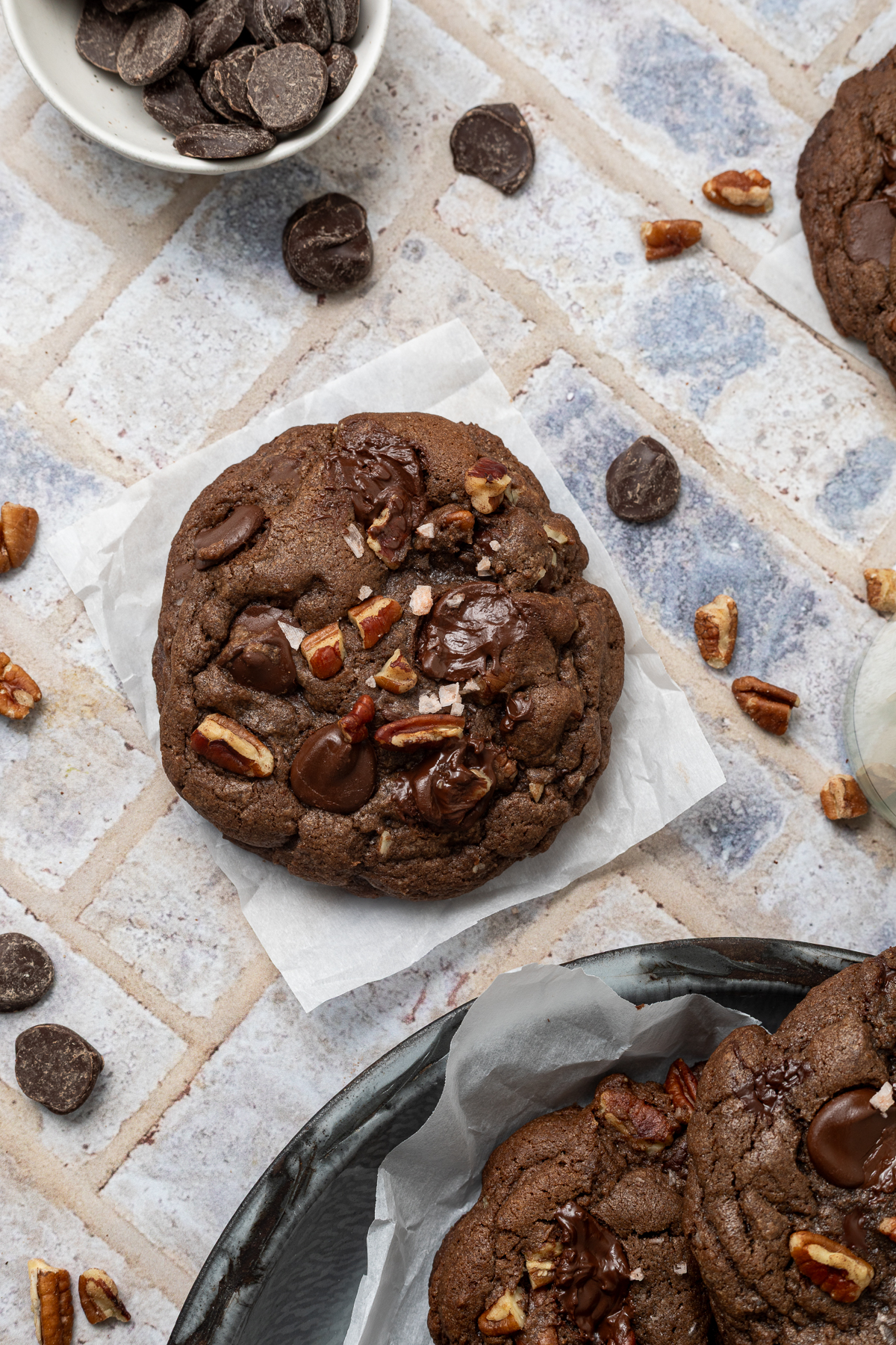 A single double chocolate chip cookie in a piece of parchment paper next to a platter of cookies and a bowl of chocolate chips.