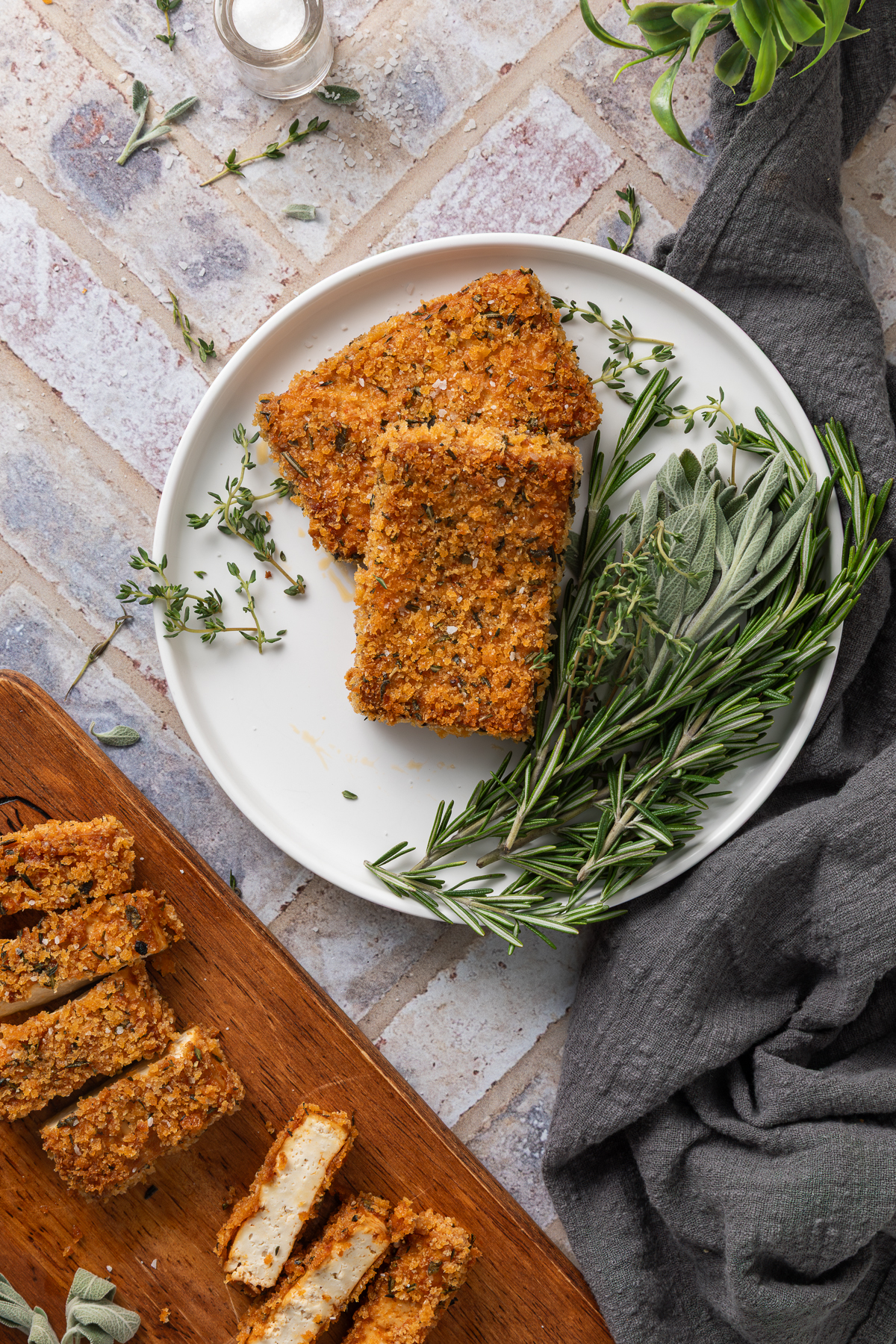 Two pieces of crispy baked tofu on a white plate next to fresh thyme, sage, and rosemary.