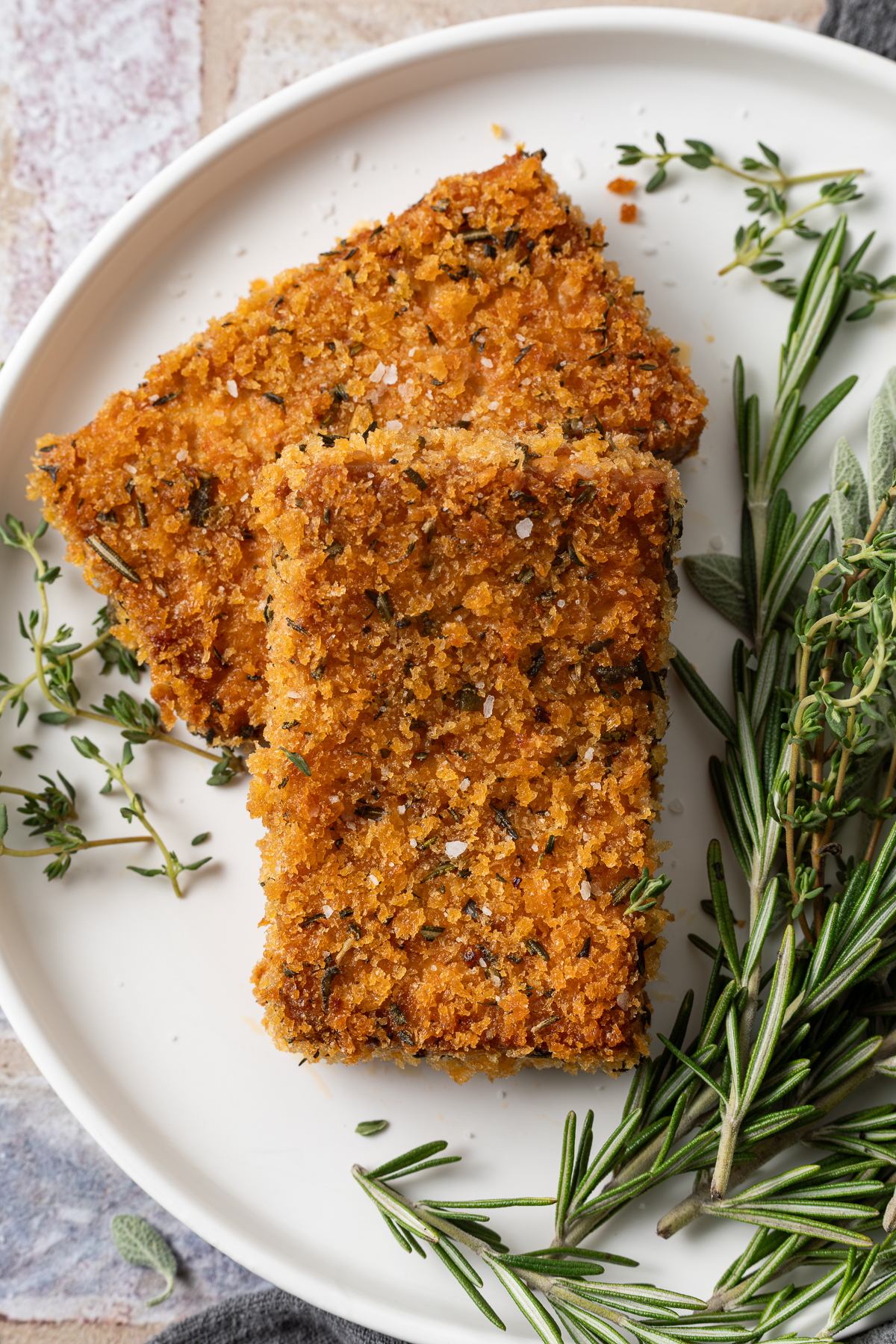 Two pieces of crispy baked tofu on a white plate next to fresh thyme, sage, and rosemary. 