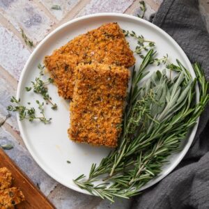 Two pieces of crispy baked tofu on a white plate next to fresh thyme, sage, and rosemary.