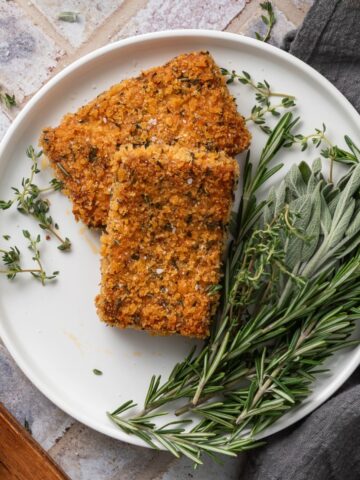 Two pieces of crispy baked tofu on a white plate next to fresh thyme, sage, and rosemary.
