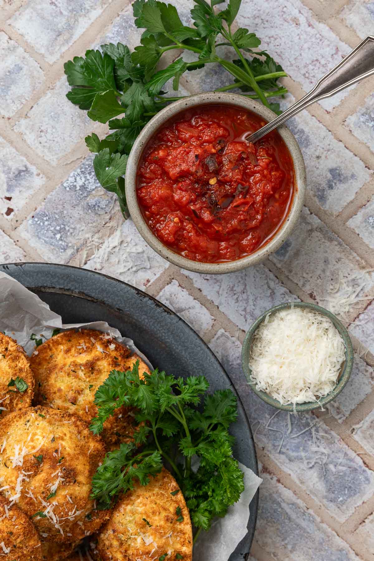 A small bowl of marinara dipping sauce with a spoon in it next to a plate of air fried ravioli and a small bowl of parmesan.