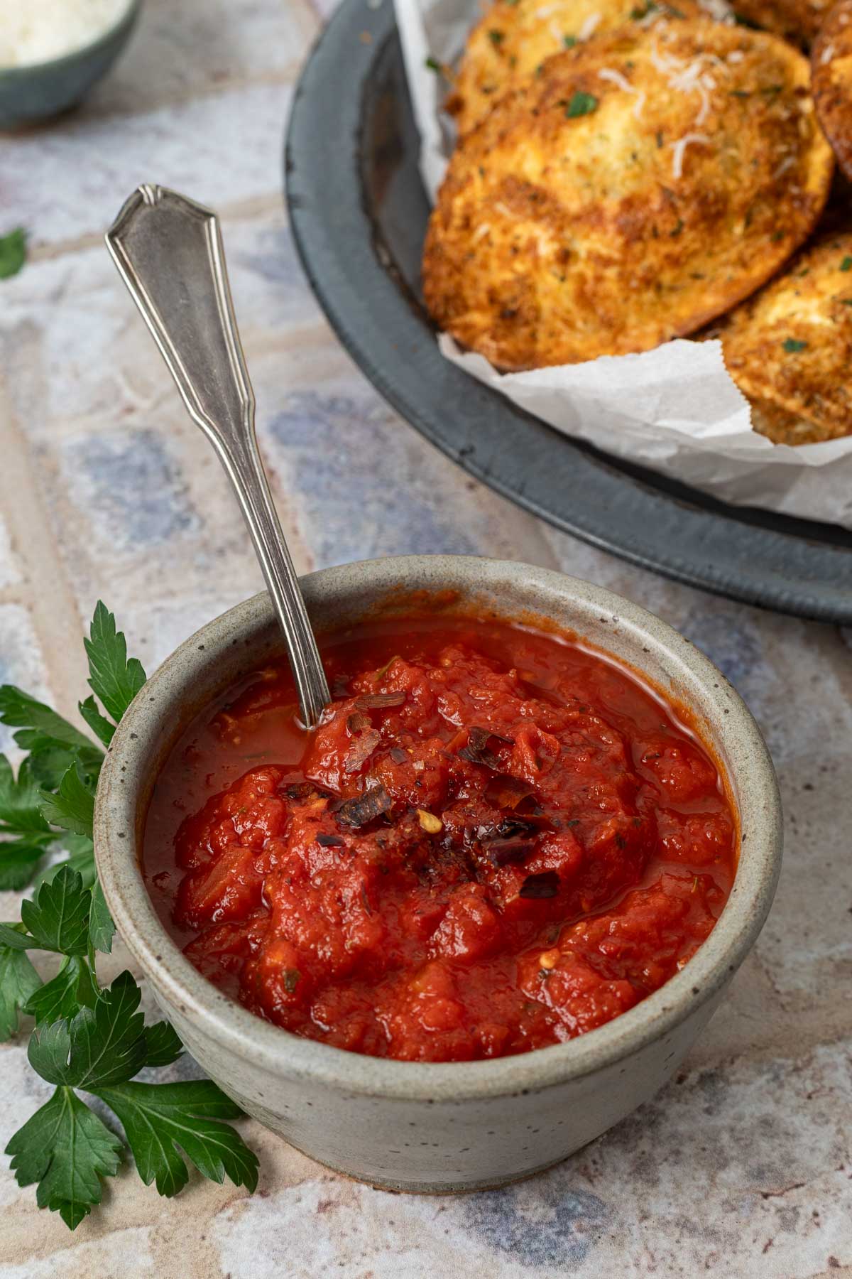 A small bowl of marinara dipping sauce with a spoon in it next to a plate of air fried ravioli.