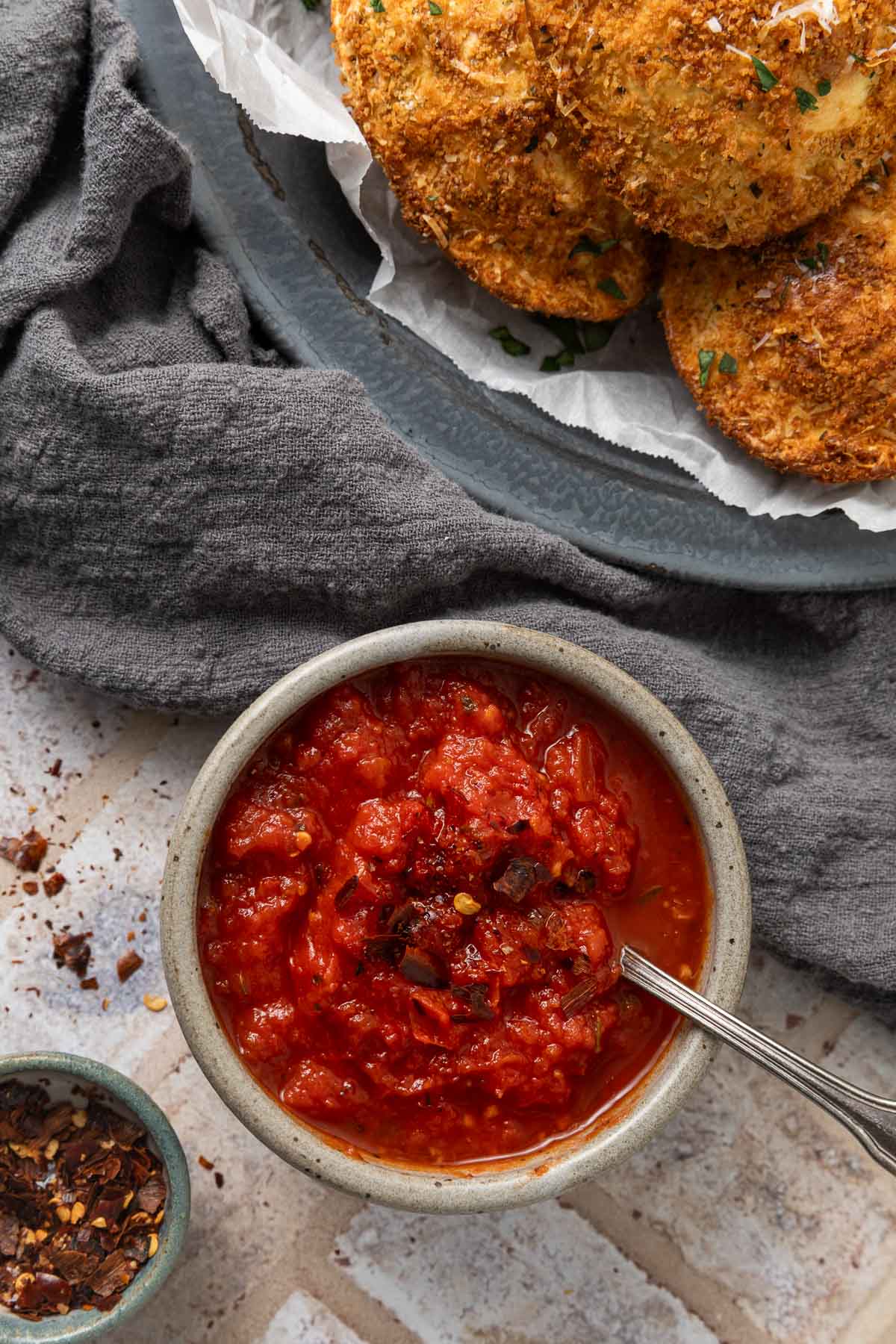 A small bowl of marinara dipping sauce with a spoon in it next to a plate of air fried ravioli.