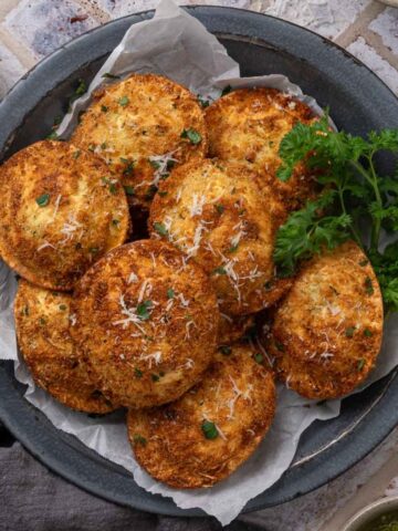 A pile of air fryer fried ravioli on a plate garnished with grated parmesan cheese and parsley.