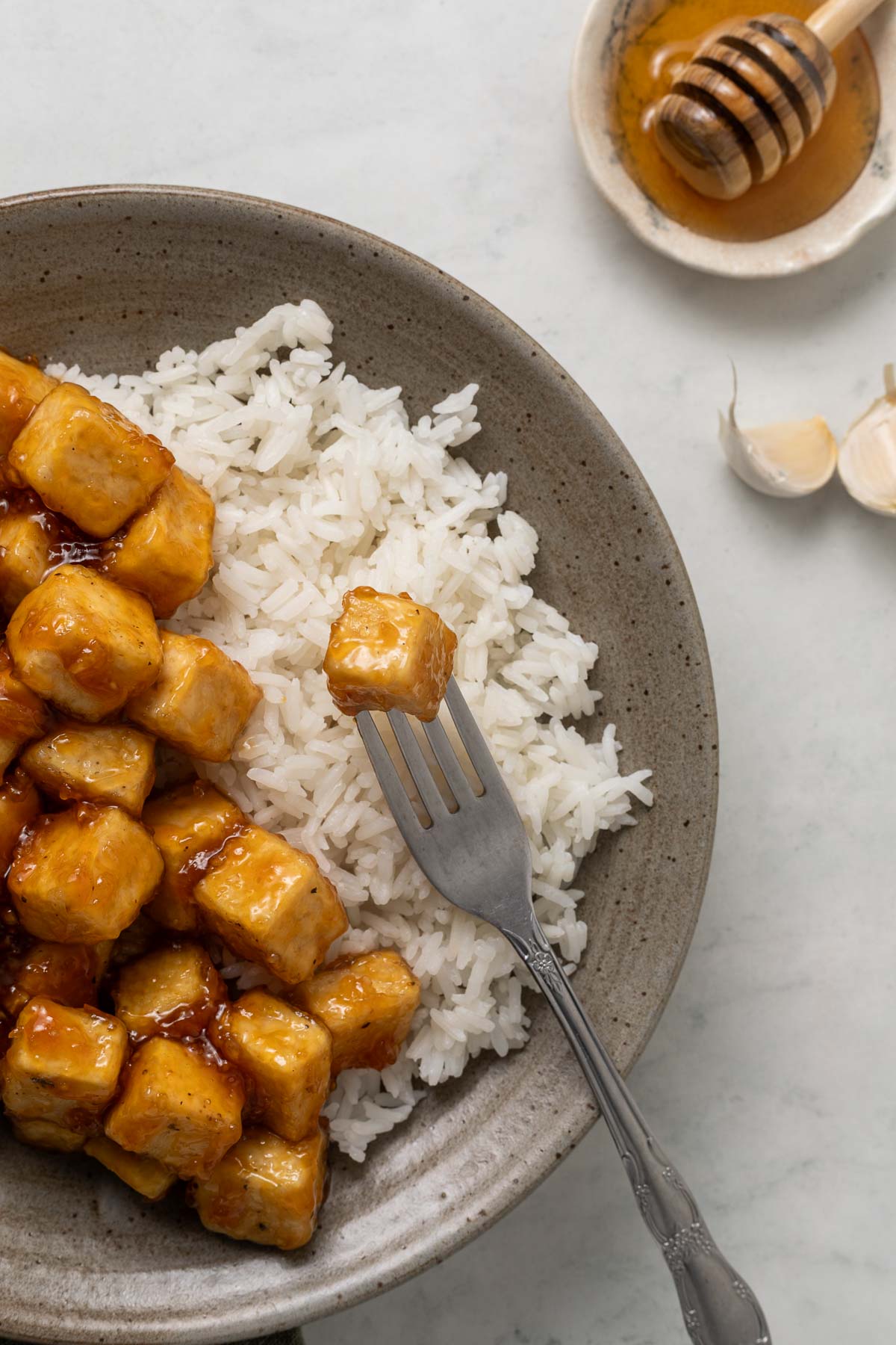 Sticky honey garlic tofu on a plate next to white rice, once piece of tofu has a fork in it.