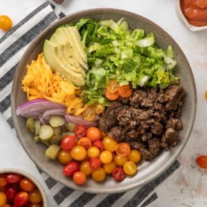 A plate of vegetarian big mac salad next to a cutting board with pickles on it, a bowl of grape tomatoes, half an avocado, and a cup of dressing.