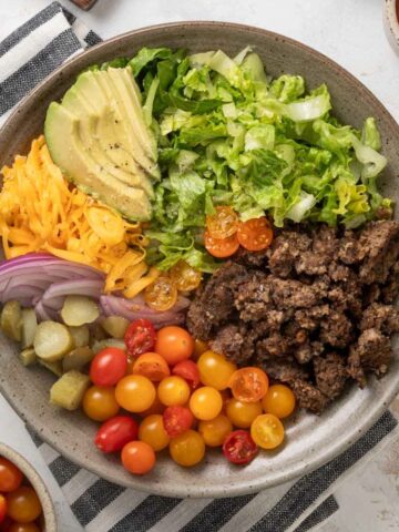 A plate of vegetarian big mac salad next to a cutting board with pickles on it, a bowl of grape tomatoes, half an avocado, and a cup of dressing.
