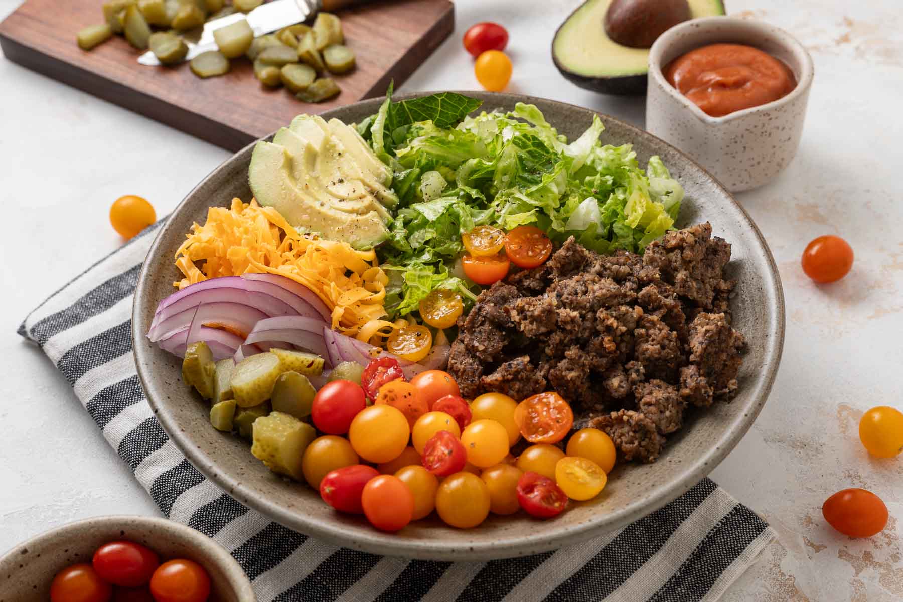 A plate of vegetarian big mac salad next to a cutting board with pickles on it, a bowl of grape tomatoes, half an avocado, and a cup of dressing.