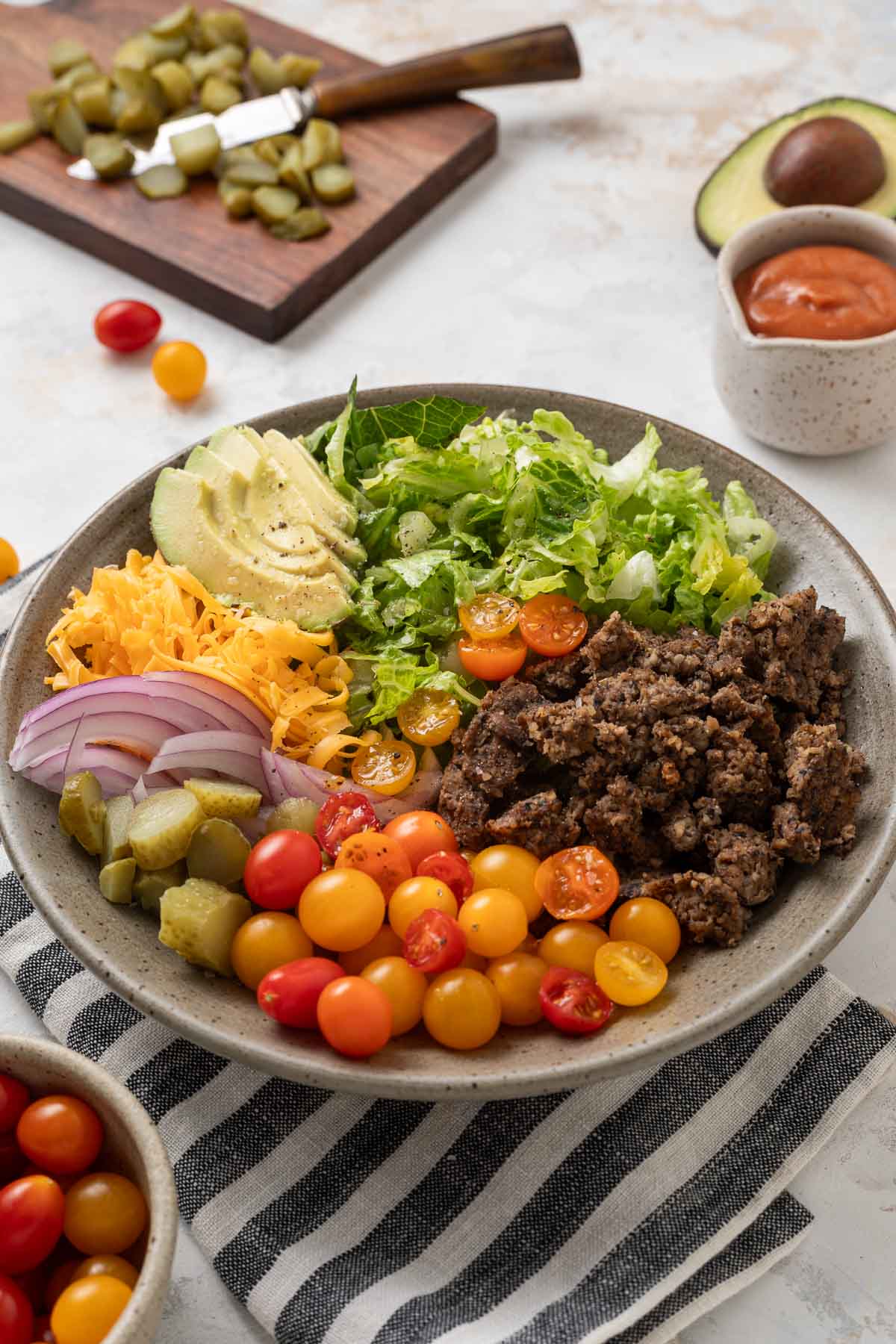 A plate of vegetarian big mac salad next to a cutting board with pickles on it, a bowl of grape tomatoes, half an avocado, and a cup of dressing.
