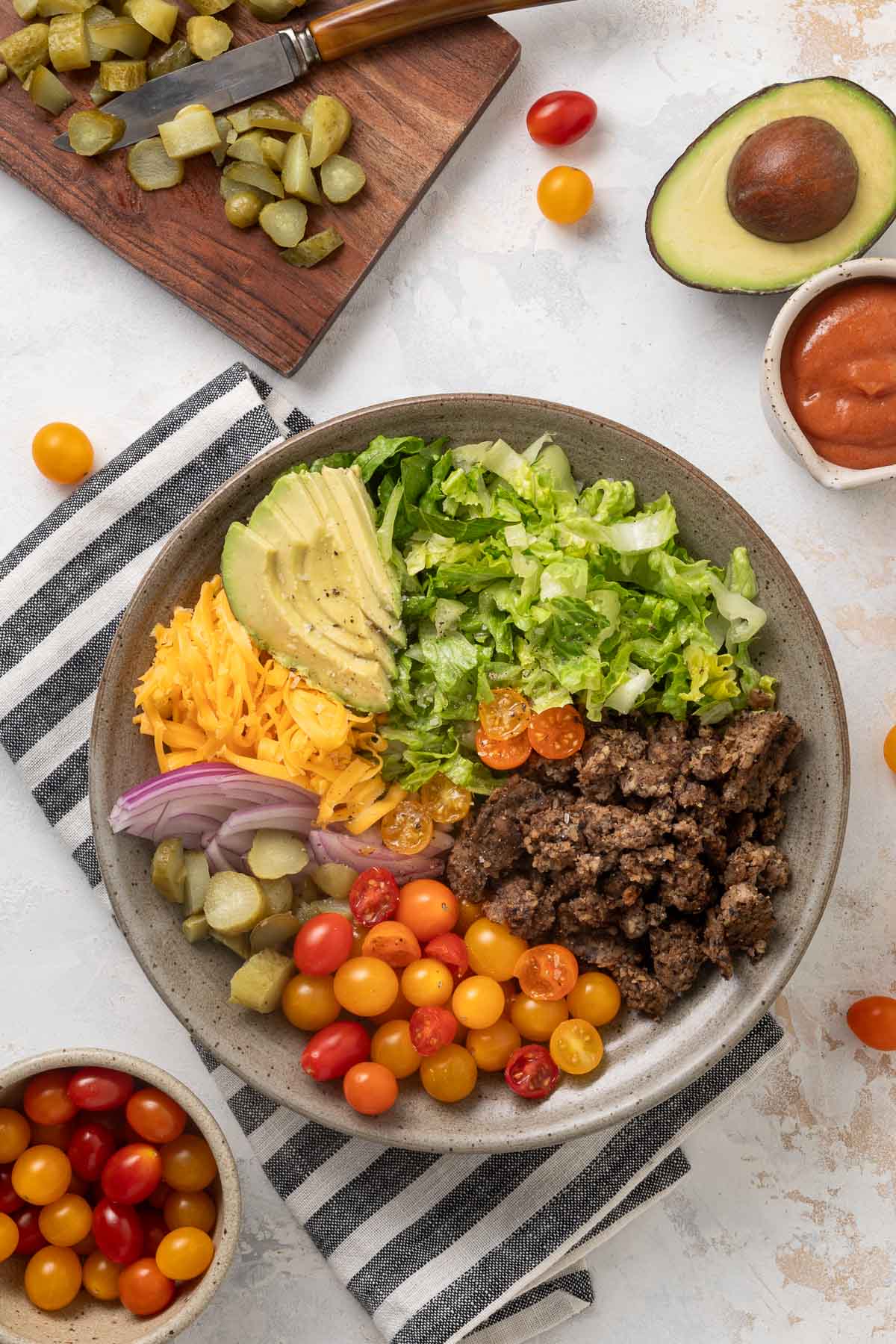 A plate of vegetarian big mac salad next to a cutting board with pickles on it, a bowl of grape tomatoes, half an avocado, and a cup of dressing.