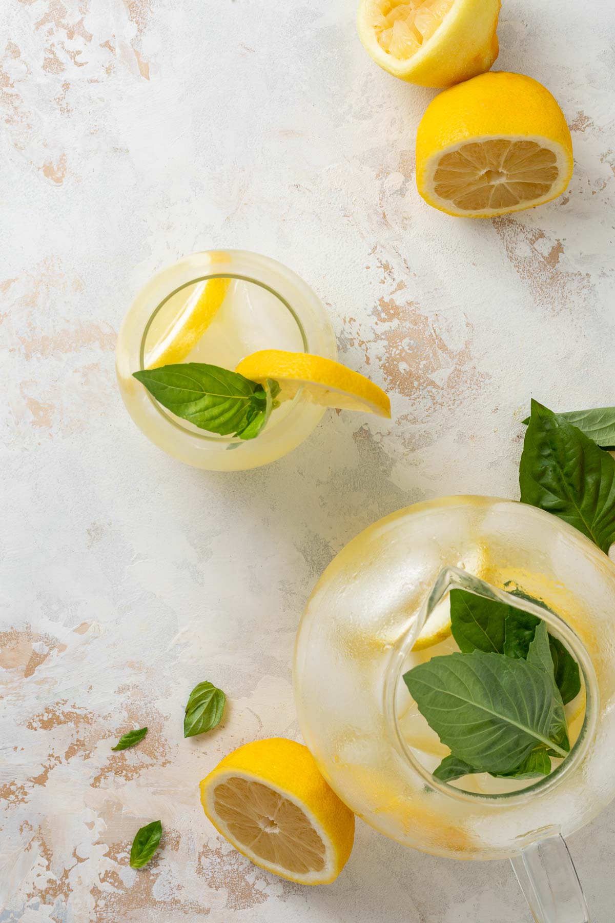 A glass of basil lemonade and a pitcher of basil lemonade garnished with fresh lemon slices and fresh basil.